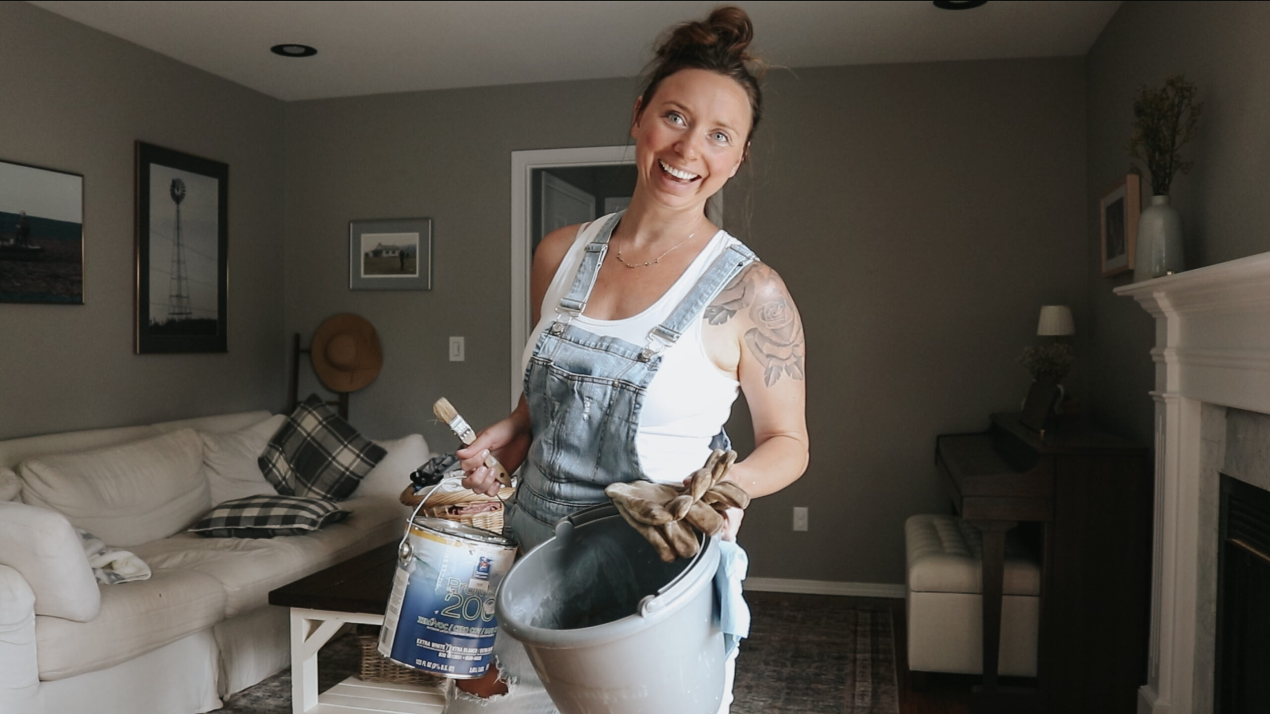 Christian homemaker wearing a white tank top and jean overalls is holding paint supplies and smiling